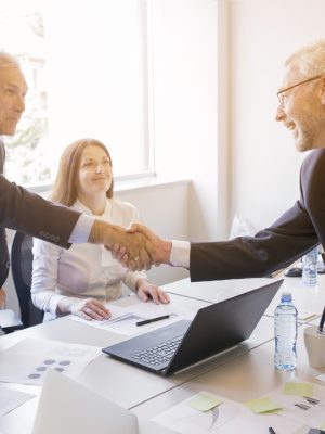smiling-woman-looking-two-senior-businessman-shaking-hands smiling-woman-looking-two-senior-businessman-shaking-hands